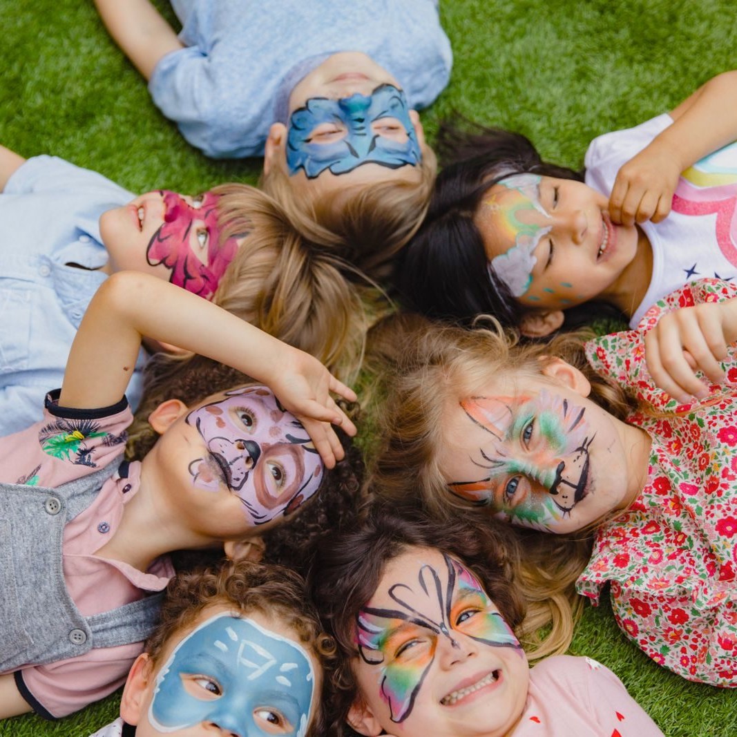 Group of children enjoying an outdoor party with colorful face paint, lying on grass.