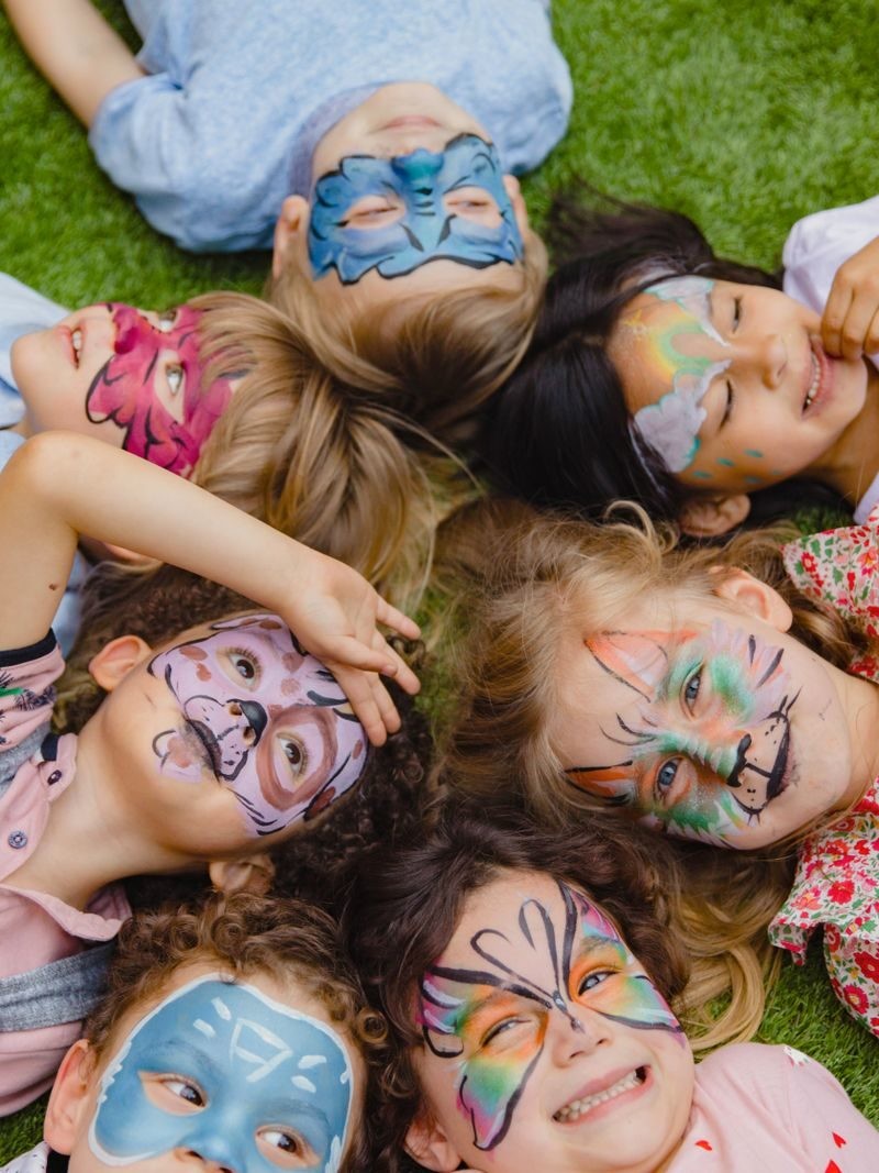 Group of children enjoying an outdoor party with colorful face paint, lying on grass.